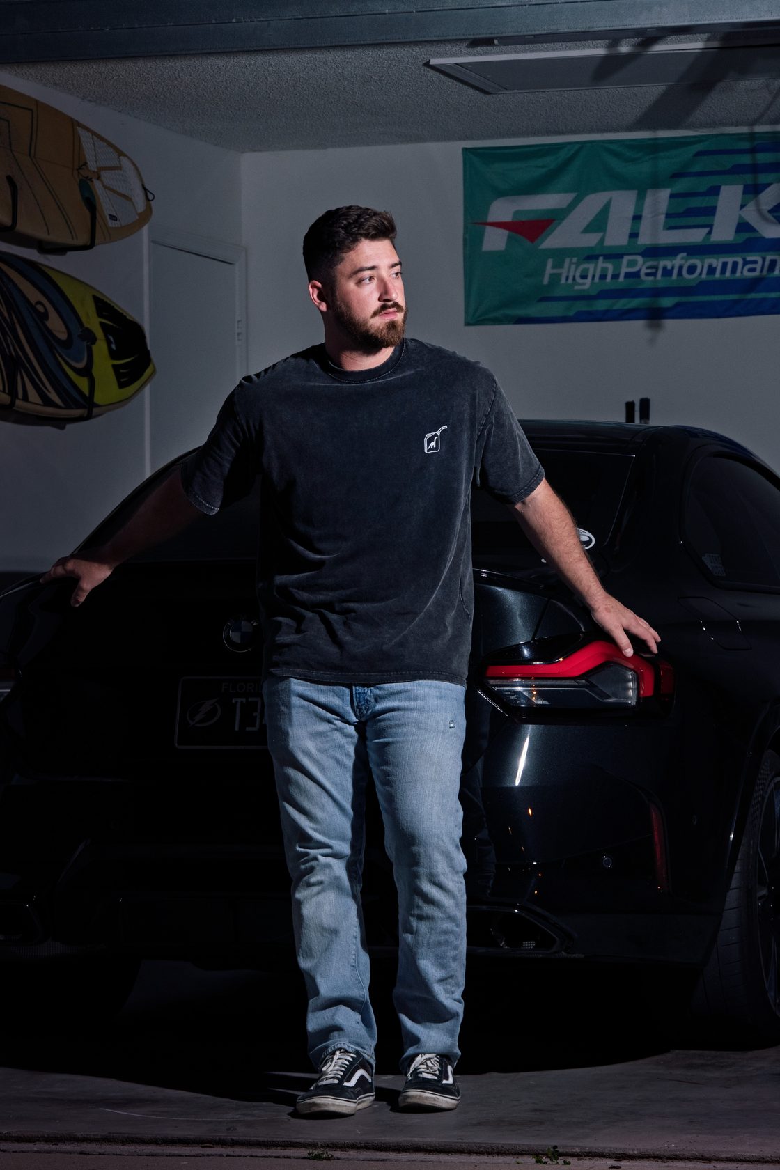 A person in a washed Dino Juice tee leaning on a BMW in the garage